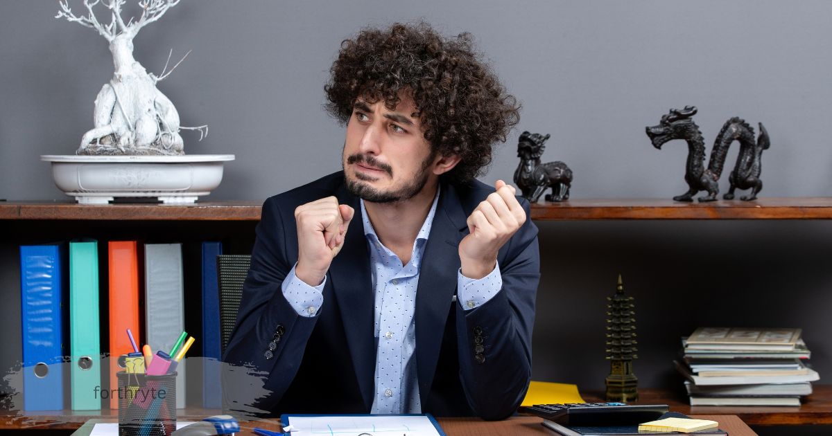 Frustrated businessman clenching fists at office desk with bookshelves and figurines, forthryte.