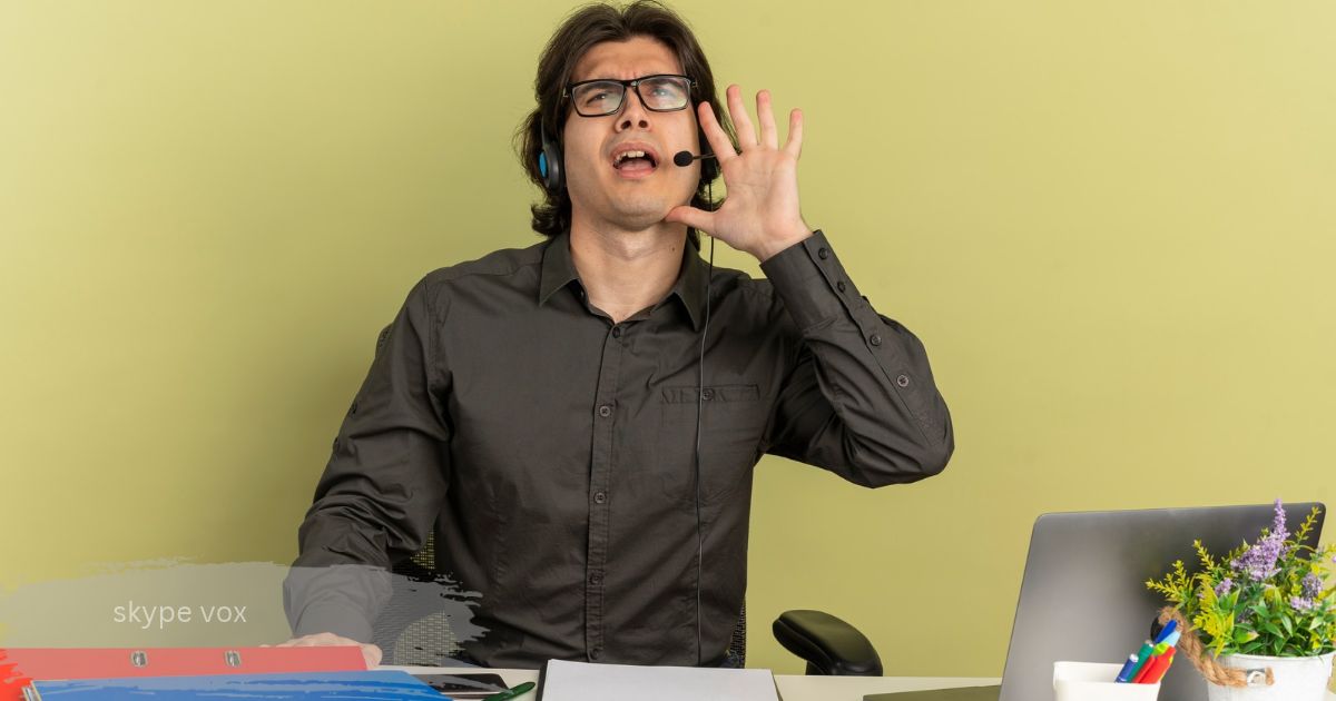 Man in glasses and a headset skype vox raising his hand while sitting at a desk with a laptop