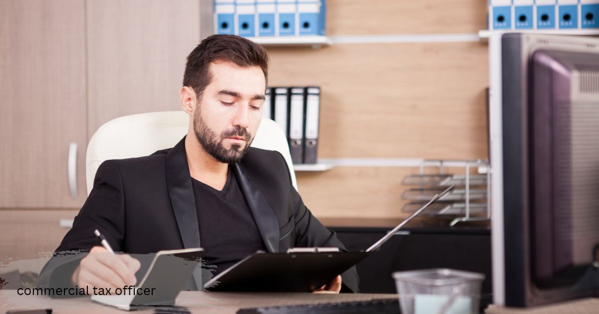Bearded professional in a black suit commercial tax officer writing notes on a clipboard at his office desk with files on the shelves behind him’commercial tax officer’