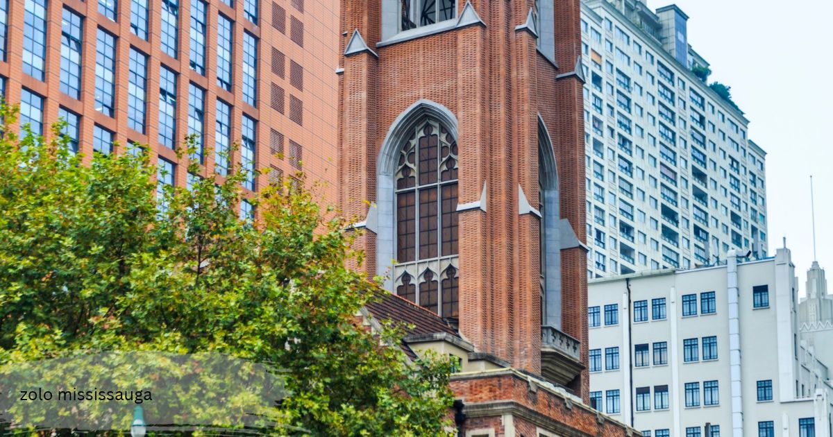 Red brick church tower with a tall Gothic arched window surrounded by green trees and modern high rise buildings zolo mississauga