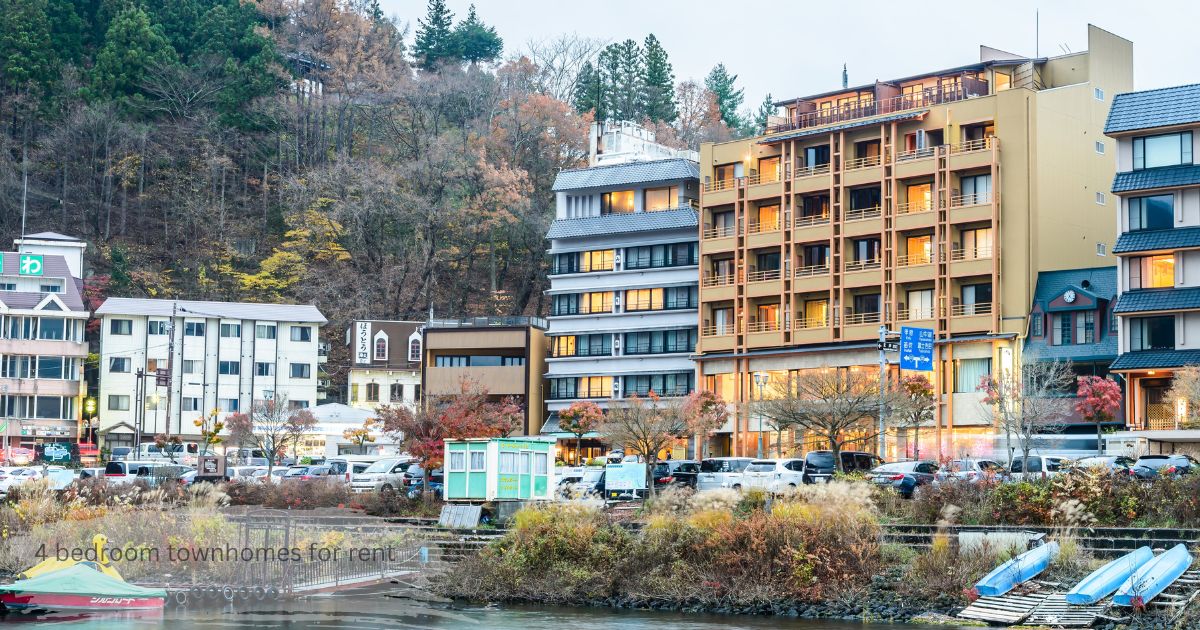 Multi story townhomes and residential buildings by a river surrounded by autumn trees, showing 4 bedroom townhomes for rent in a scenic town setting.