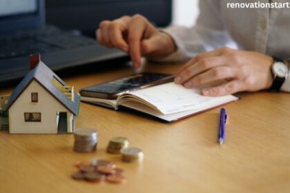 Person calculating cost of home renovation with calculator, notebook, small house model, and stacked coins on desk