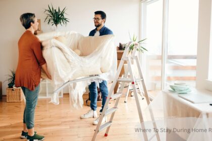 Couple wrapping furniture in plastic while packing up their home during a renovation, with moving boxes and a ladder in the background — where to stay during home renovation.