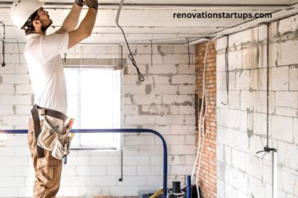 Construction worker doing an apartment renovation, wearing a hard hat and tool belt while wiring the ceiling on a scaffold, surrounded by exposed brick, concrete block walls, and hanging electrical cables in an unfinished room.