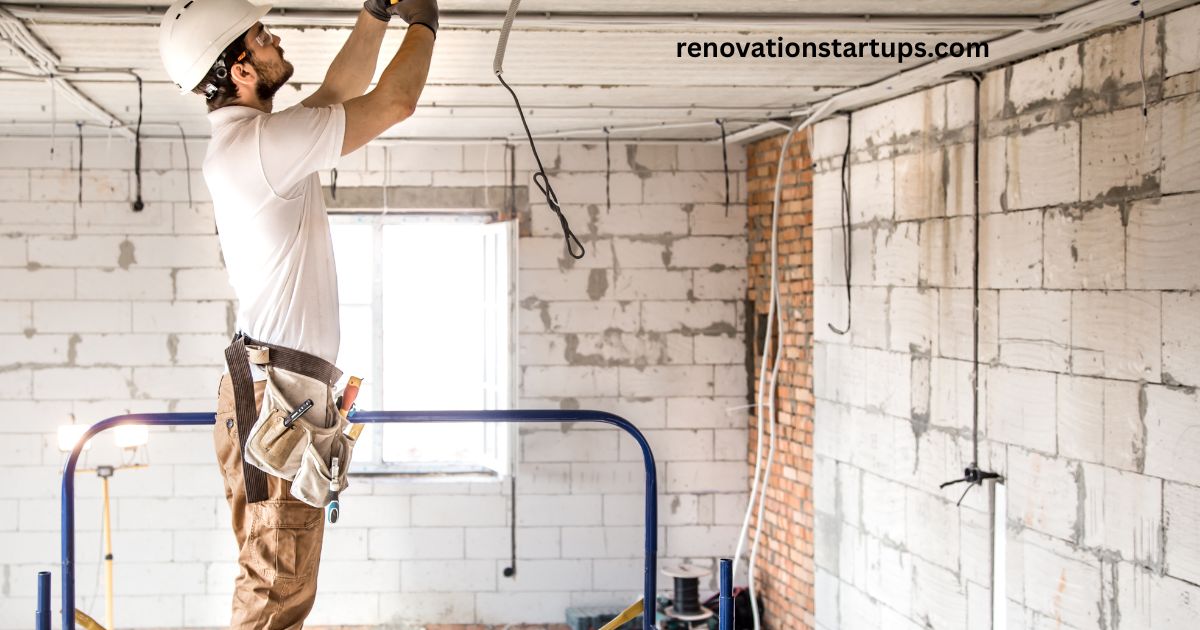 Construction worker doing an apartment renovation, wearing a hard hat and tool belt while wiring the ceiling on a scaffold, surrounded by exposed brick, concrete block walls, and hanging electrical cables in an unfinished room.