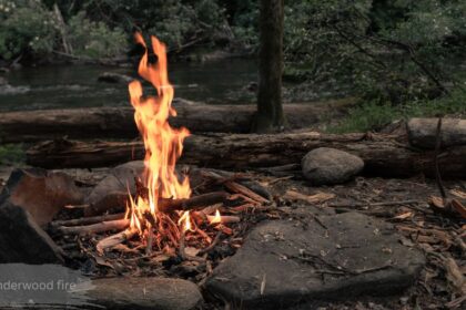 Woman warming her hands over a small campfire while holding a red mug, forested hillside in the background — mount underwood fire