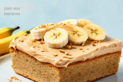 A slice of banana cake with smooth icing, topped with fresh banana slices on a white plate’ Pain Aux Bananes’.