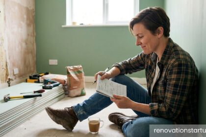 = A homeowner sitting on the floor of an unfinished room next to drywall, reviewing a renovation checklist to figure out how to stop procrastinating on home improvement projects.