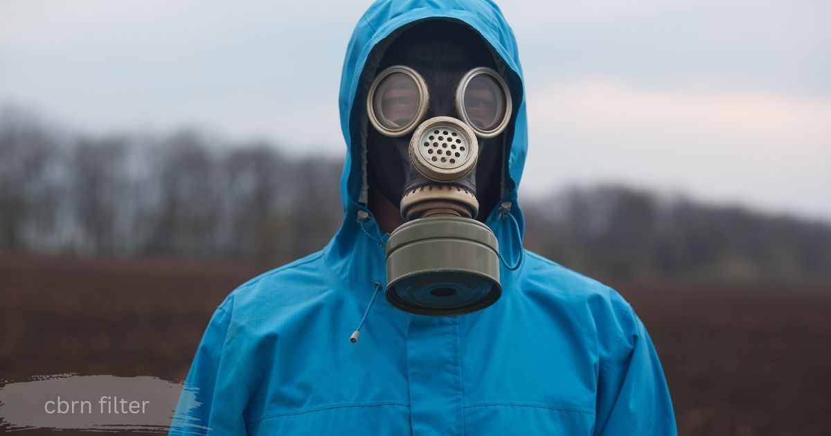 Person in a white hooded coverall wearing a black respirator mask with a yellow filter cartridge, examining a small dried plant specimen held in tweezers over a glass vial, outdoors near a rocky shoreline.