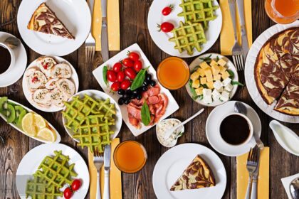 Brunch spread on wooden table with matcha waffles, coffee, cheesecake, and fresh fruit.