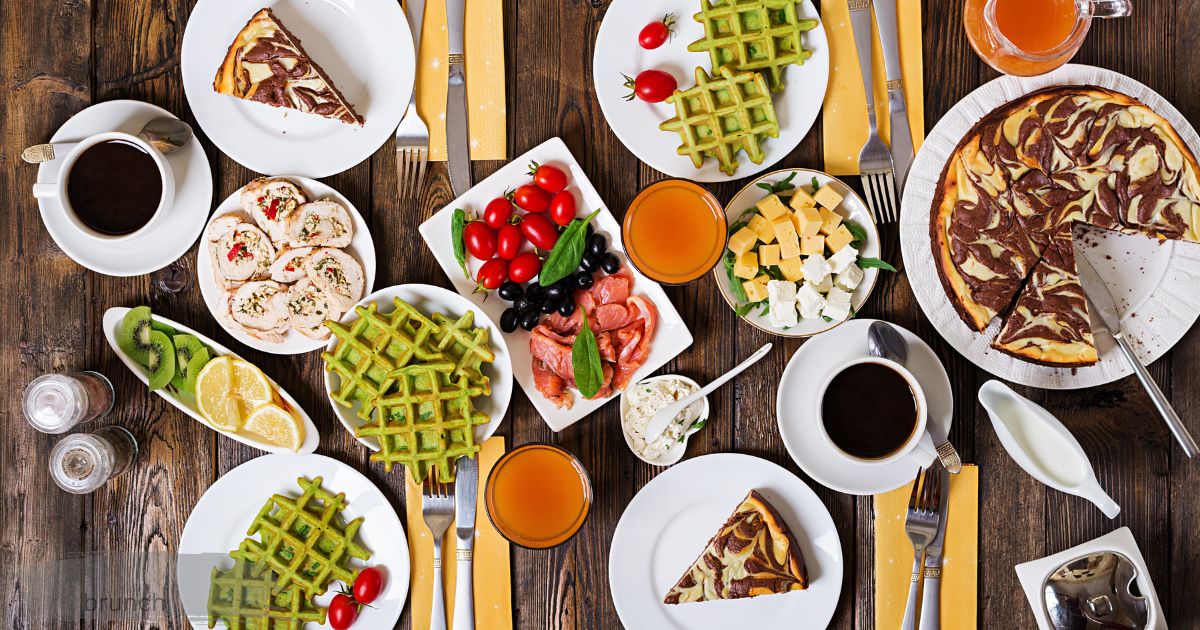 Brunch spread on wooden table with matcha waffles, coffee, cheesecake, and fresh fruit.