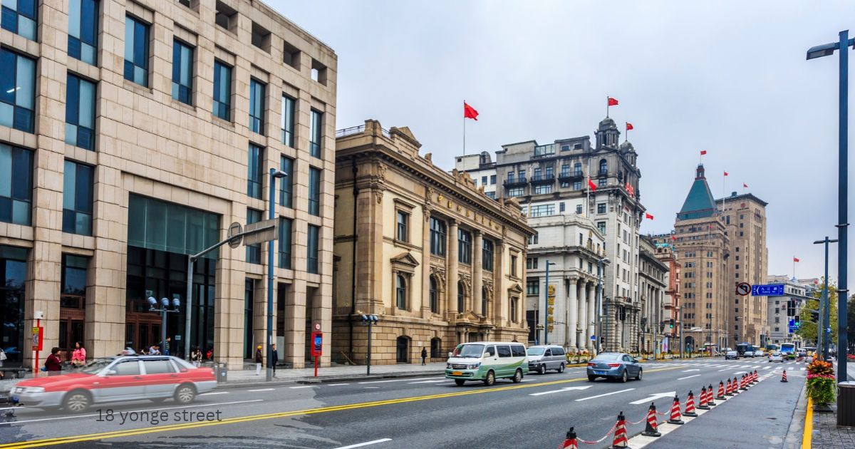 Historic stone buildings and a modern glass building along a city street with cars, red flags and traffic cones, 18 yonge street